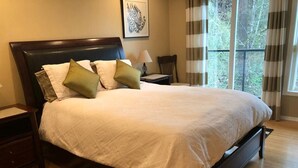 A neatly made bed with a white quilt, a dark headboard, and two green pillows. There is a wooden nightstand with a lamp beside the bed, and a chair in the corner. A large window with curtains is visible in the background. - Zen themed private retreat in NW Portland. (Portland)