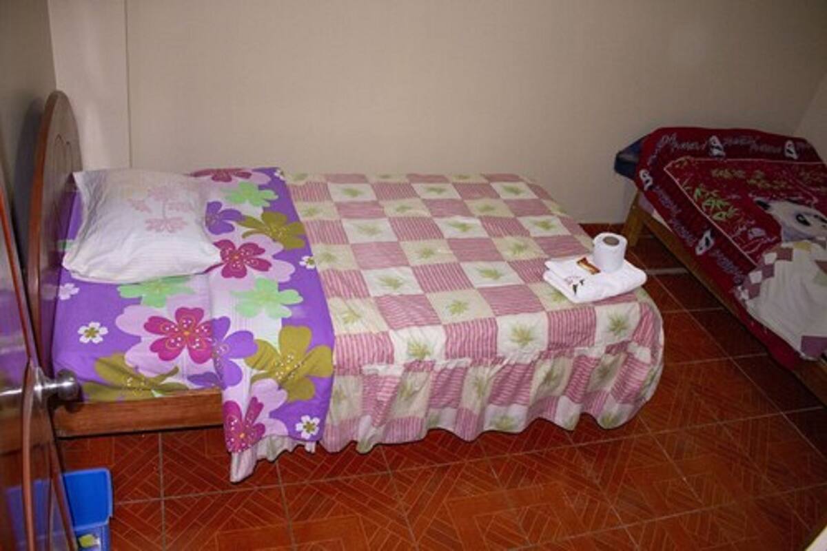 Une petite chambre avec un lit simple, recouverte d’une courtepointe à motifs floraux, d’une couverture à carreaux et d’un rouleau de papier toilette posé sur le lit.