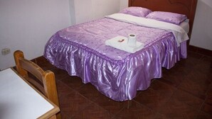 A bedroom with a bed covered in purple bedding, a wooden headboard, and a small table with a chair. - Hospedaje Ollantays (Cusco)