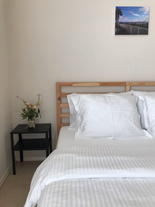 A bedroom with a wooden headboard, a bed with white bedding, a small black nightstand with a vase of flowers, and a framed picture on the wall.