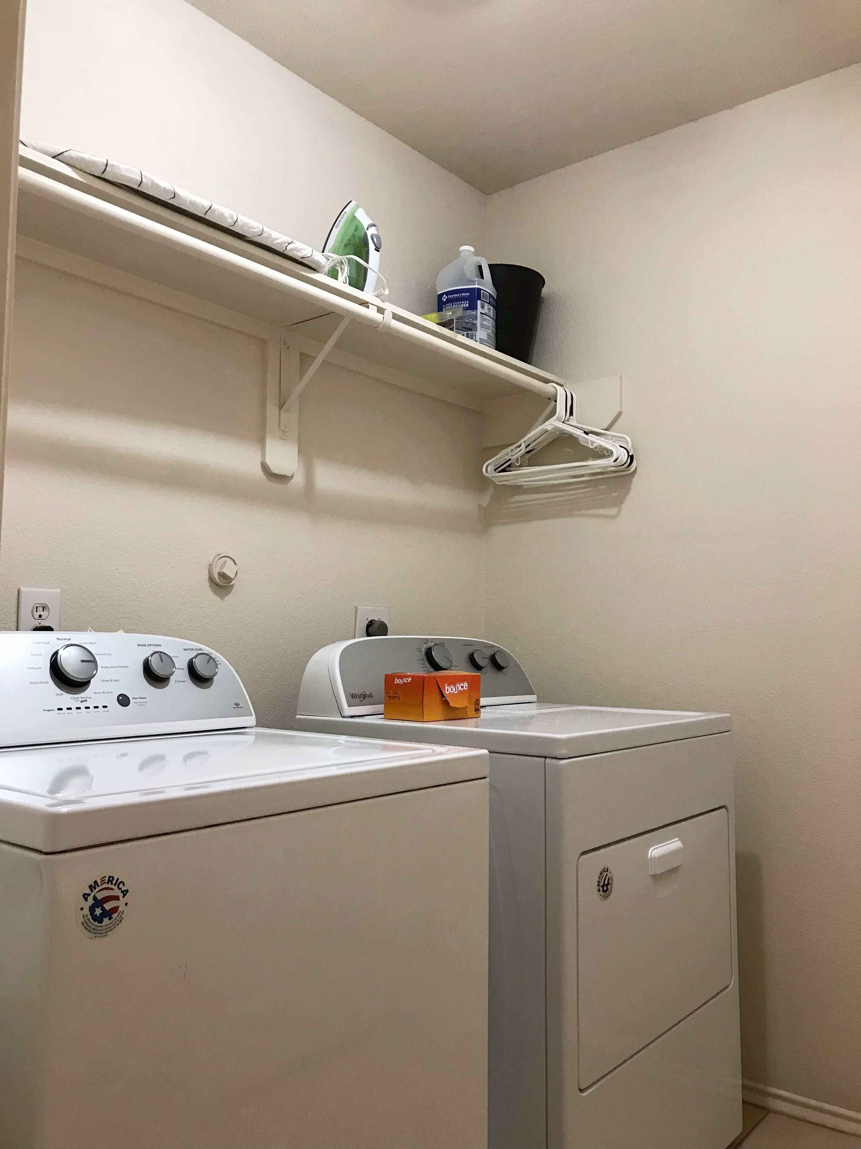A laundry area with a washing machine and dryer, a shelf with a bottle of detergent and an iron, and a wall-mounted outlet.