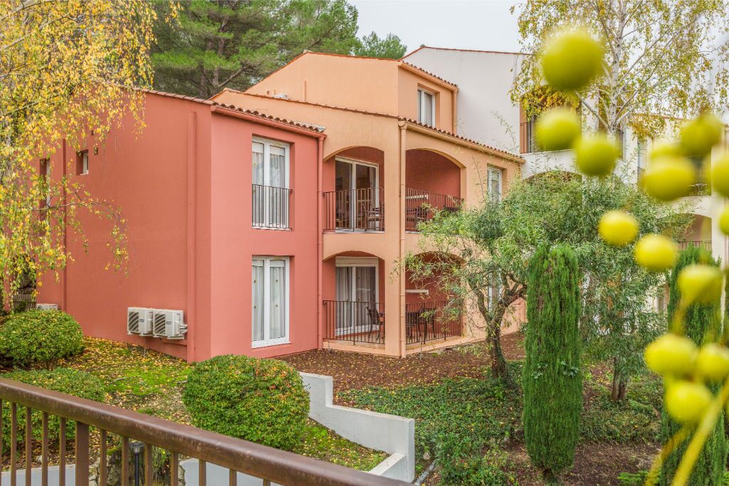 A two-story building with a red facade, balconies, and a garden area with trees and shrubs.