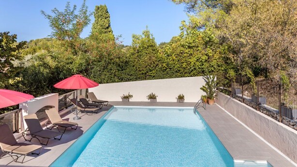 A rectangular swimming pool with lounge chairs and red umbrellas, surrounded by greenery and a stone wall.