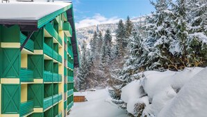 A multi-story building with green and yellow exterior walls, surrounded by snow-covered trees and a mountainous backdrop. - 1-Bed Apartment | Schladming Ski Resort | Sleeps 4 (Schladming)
