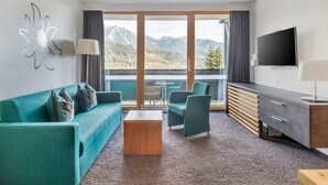 A modern hotel room with a sofa, chairs, a TV, and a balcony with mountain views. - 1-Bed Apartment | Schladming Ski Resort | Sleeps 4 (Schladming)