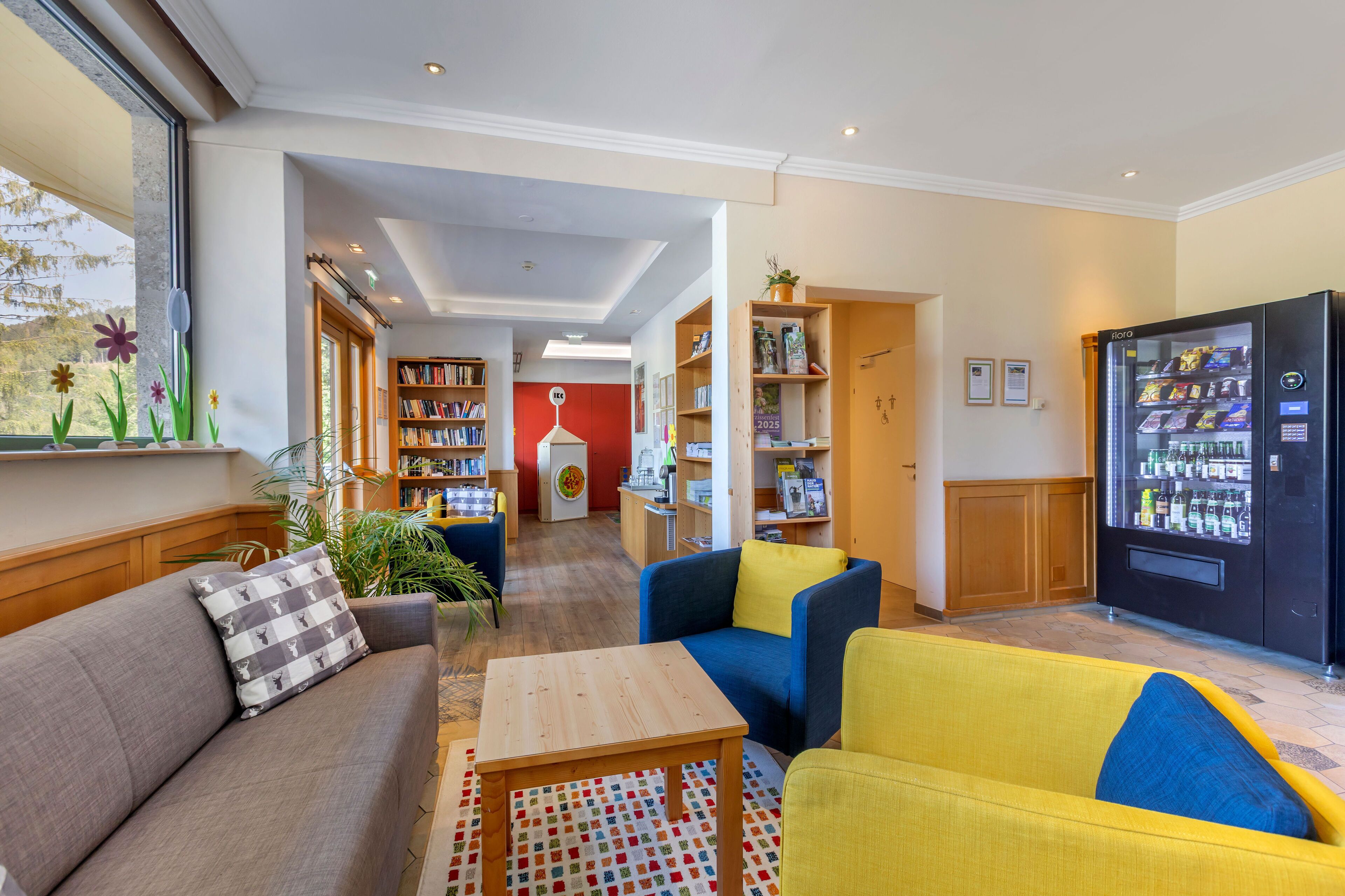 Un hall d’hôtel avec un canapé, des fauteuils, une table basse, un réfrigérateur et une bibliothèque.