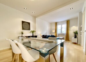 Un salon moderne avec une table à manger en verre, des chaises blanches, un canapé et une lampe.