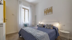 A bedroom with a bed, a bedside table, a lamp, a window with curtains, a radiator, and a wall art piece. - GuestHost - Elegant and modern 60sqm apartment that can accommodate up to 4 people. Located on the third floor, with elevator (not suitable for disabled people), of a characteristic building in San Lorenzo area, the apartment is about 15 minutes walk fro (Roma)