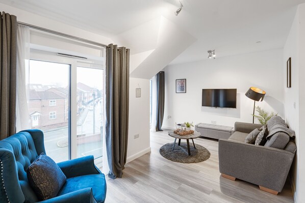 A modern living room with a blue sofa, a grey sofa, a flat-screen TV, and a large window with curtains.