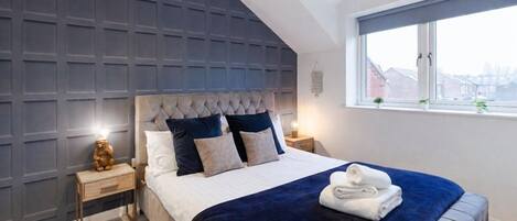 A bedroom with a tufted headboard, a large window, and a neatly made bed with a blue blanket.