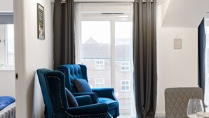 Un salon moderne avec un canapé bleu, une table à manger dressée pour deux personnes et une vue sur un bâtiment en briques par la fenêtre.