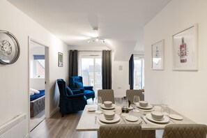 A dining area with a table set for four, a blue armchair, and a view through a window. - Aura Retreat- 1 Bedroom Apartment (York)