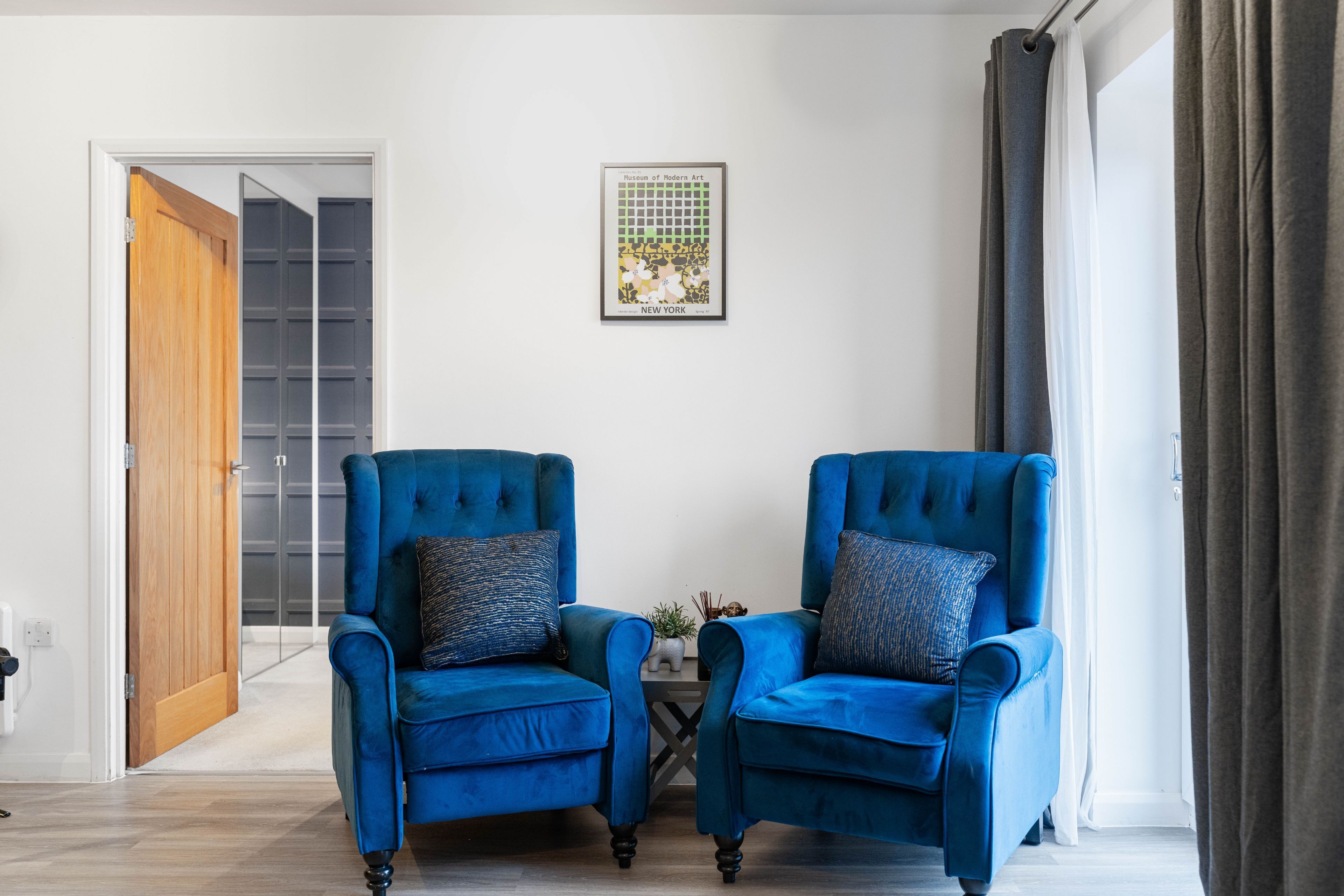 A room with two blue armchairs, a small table, a framed picture on the wall, and a door leading to another room.