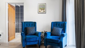 Une pièce avec deux fauteuils bleus, une petite table, un tableau encadré au mur et une porte donnant sur une autre pièce.