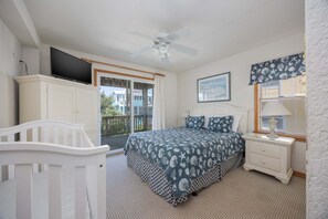 A bedroom with a bed, a nightstand, a TV, a ceiling fan, and a window with a view of buildings. - 1818 - It's A Shore Thing, 70 Yards to Beach | Pool, Hot Tub & Elevator (Corolla)