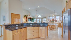 A spacious kitchen with a granite countertop, wooden cabinets, and a large island. There is a dining area with a table and chairs, and a refrigerator on the right. - 1818 - It's A Shore Thing, 70 Yards to Beach | Pool, Hot Tub & Elevator (Corolla)