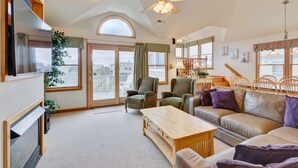 A spacious living room with a large sofa, a wooden coffee table, and two armchairs. There is a ceiling fan, a TV mounted on the wall, and a large window with a view of the city. - 1818 - It's A Shore Thing, 70 Yards to Beach | Pool, Hot Tub & Elevator (Corolla)