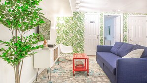 A living room with a blue sofa, a white desk, a small red cart, and a large green leafy plant.
