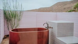 A large copper bathtub with a modern faucet, set against a backdrop of a desert landscape and a corrugated metal wall.