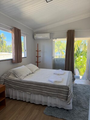 Een slaapkamer met een groot bed, een houten nachtkastje, een raam met gordijnen en uitzicht op palmbomen.