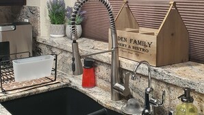 A kitchen with a black sink, stainless steel faucet, and granite countertop. There are decorative items like a potted plant and a wooden box with text.