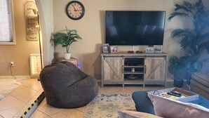 A living room with a large flat-screen TV, a bean bag chair, a potted plant, and a small cabinet with decorative items.