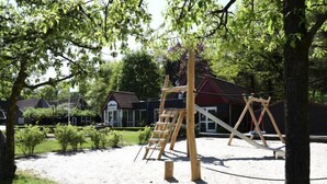 Un terrain de jeux avec des balançoires et une structure d’escalade, entouré d’arbres et avec un bâtiment en arrière-plan.
