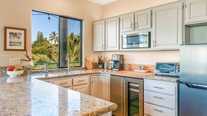 A modern kitchen with granite countertops, stainless steel appliances, and a large window offering a view of palm trees. - Spectacular Panoramic Ocean Views - Private Ohana - Wailea Custom Pole Home (Kihei)