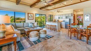 A spacious living area with a sofa, coffee table, and ottomans, a dining area with a table and chairs, and a kitchen with cabinets and appliances. - Spectacular Panoramic Ocean Views - Private Ohana - Wailea Custom Pole Home (Kihei)