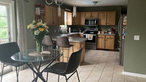 A modern kitchen with wooden cabinets, a central island, and a dining area with a glass table and chairs. - Crew Road Cabana (Clifton Park)