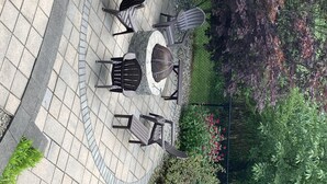 A patio area with a round table and four chairs, surrounded by a stone floor and bordered by a fence with greenery and flowers. - Crew Road Cabana (Clifton Park)