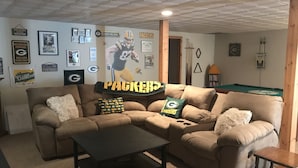 A living room with a sectional sofa, a coffee table, and a pool table. The walls are decorated with sports memorabilia and a large poster of a football player. - Crew Road Cabana (Clifton Park)