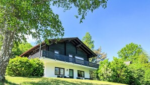 Un chalet de dos pisos con un balcón, rodeado de exuberante vegetación y árboles.