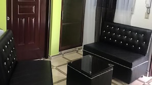 Una habitación con una puerta de madera oscura, pared verde, sofá de cuero negro, mesa de centro de vidrio y cortina blanca.