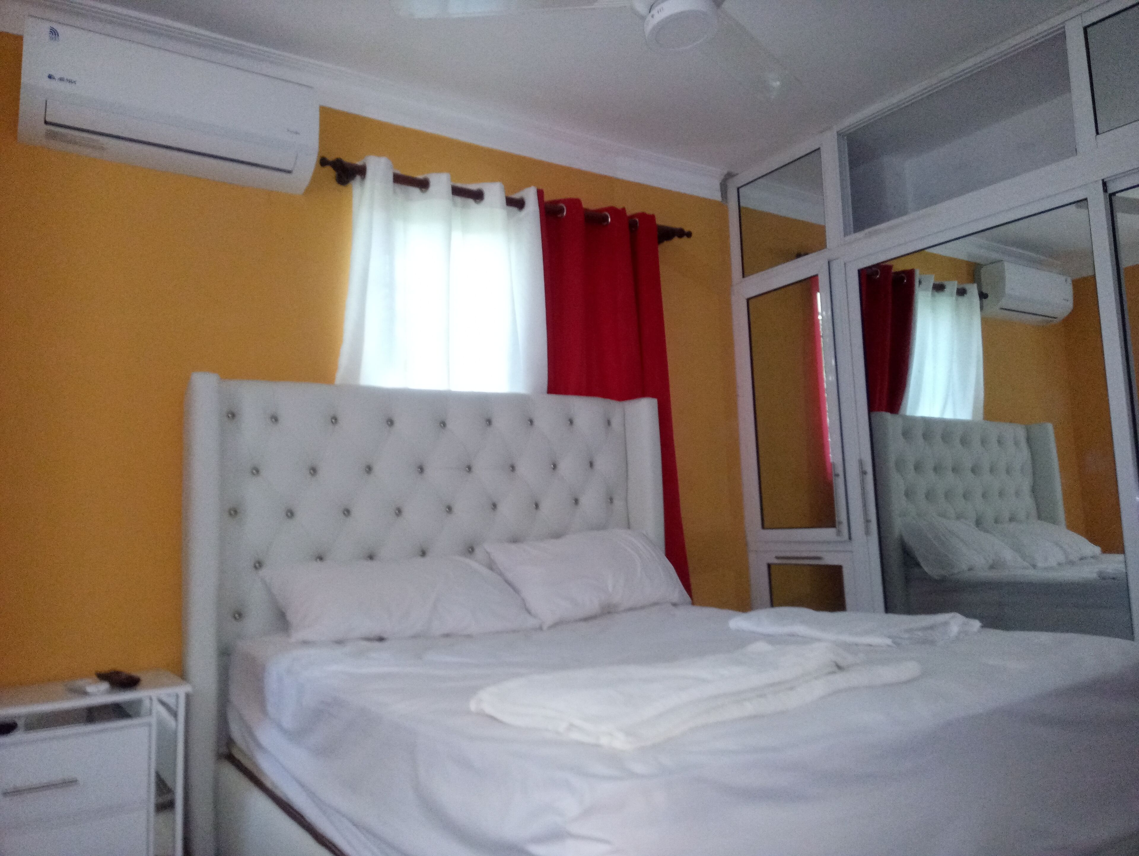 Un dormitorio con cabecera tapizada de blanco, una cama individual, una mesita de noche, un espejo y una ventana con cortinas rojas.