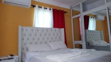 Quarto com cabeceira estofada branca, uma cama de solteiro, mesa de cabeceira, espelho e janela com cortinas vermelhas.