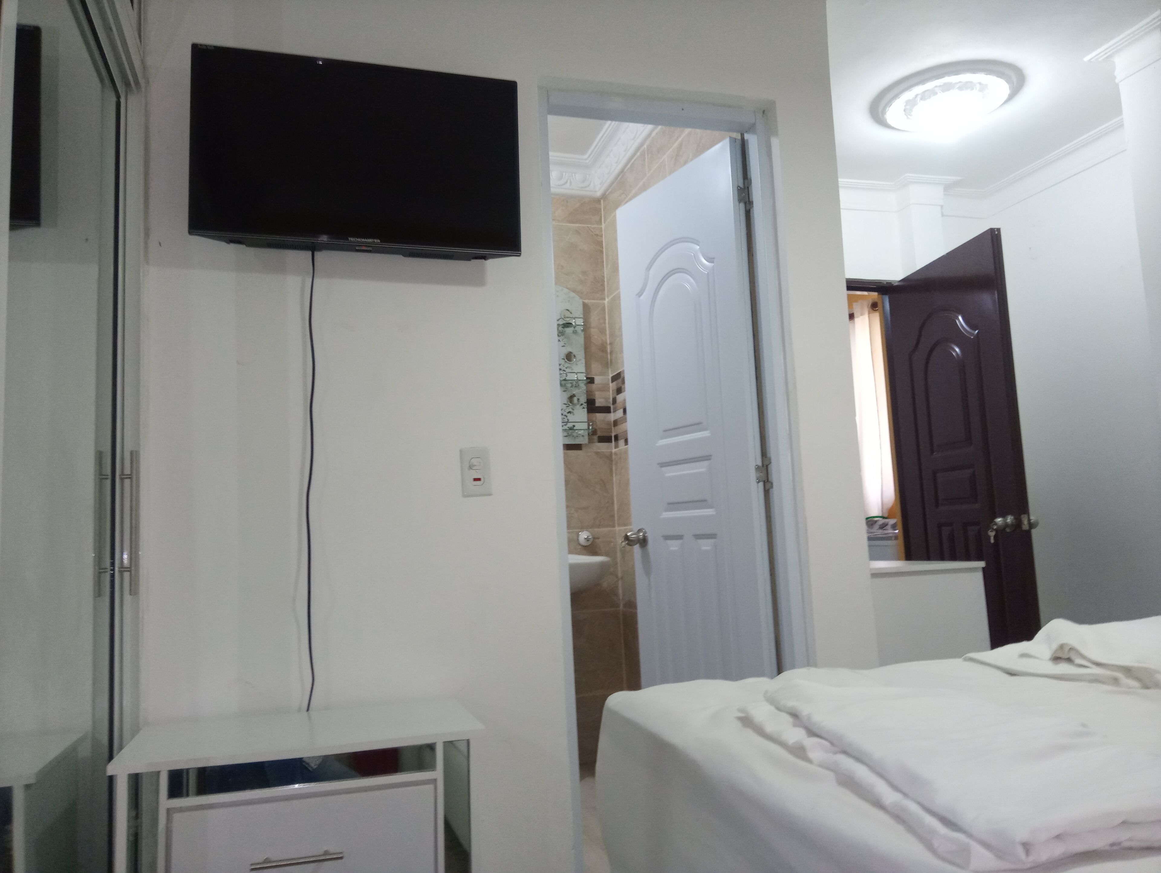 A bedroom with a bed, a TV mounted on the wall, a bedside table, and a door leading to another room.