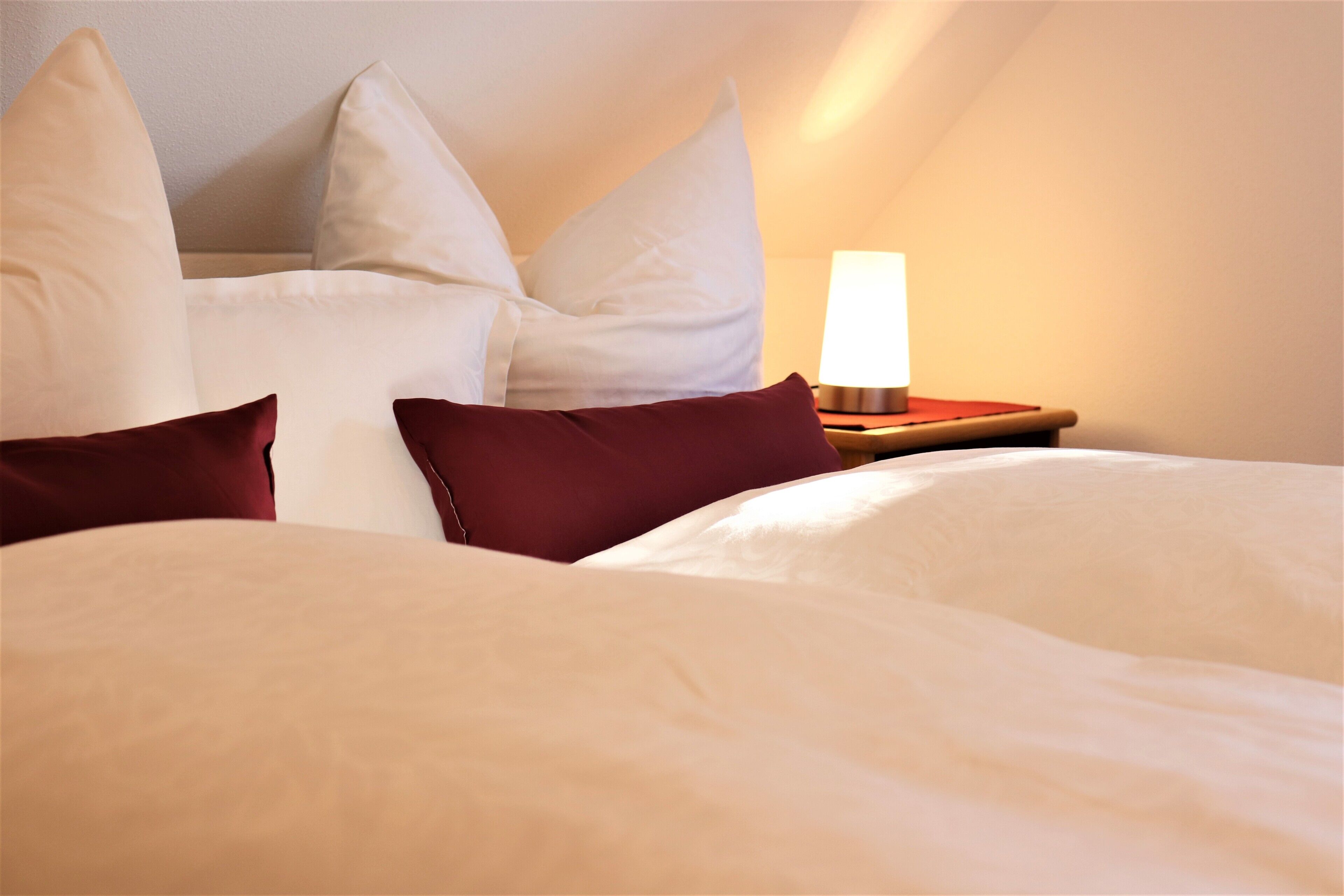 Ein ordentlich bezogenes Bett mit weißen und dunkelroten Kissen, eine Nachttischlampe und ein Holfnachtkästchen.
