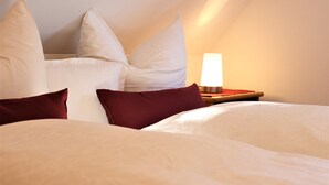 Una cama bien tendida con almohadas blancas y burdeos, una lámpara de noche y una mesita de madera.
