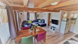 Una sala moderna con techo de madera, un sofá modular, un televisor y un comedor con mesa de madera y sillas coloridas.