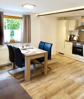 A modern kitchen with a dining area, wooden flooring, and a view of greenery outside.