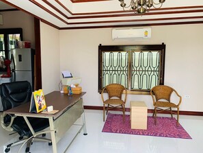 A room with a desk, two wicker chairs, a small table, a standing fan, a refrigerator, and a window with decorative metal bars.
