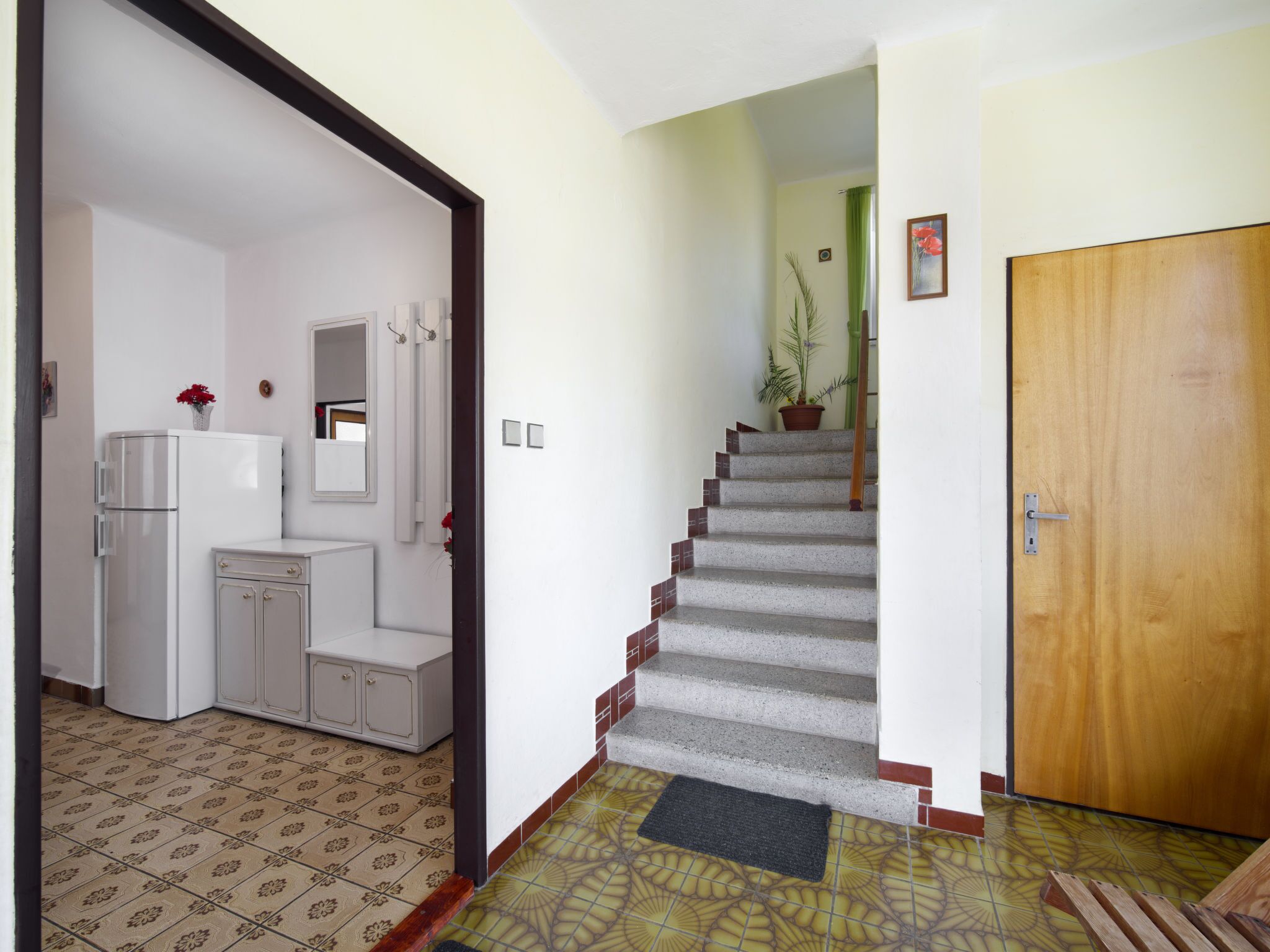 A hallway leading to a bedroom with a wooden door, a staircase, a bathroom with a toilet and sink, and a refrigerator.