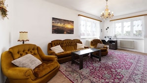 A living room with a large sofa set, a coffee table, and a patterned rug. There is a painting on the wall, a chandelier, and a window with curtains. - Neurazy by Interhome (Neurazy)