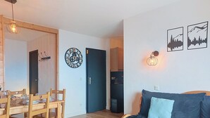 A living room with a sofa, a dining table with chairs, a clock, and wall-mounted artwork. - Nice apartment for 6 guests with WIFI, pool, TV and balcony (Le Corbier)