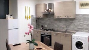 A modern kitchen with a dining area, featuring a wooden table, chairs, a refrigerator, a stove, and a washing machine. - Graphite House 2.0 (Napoli)