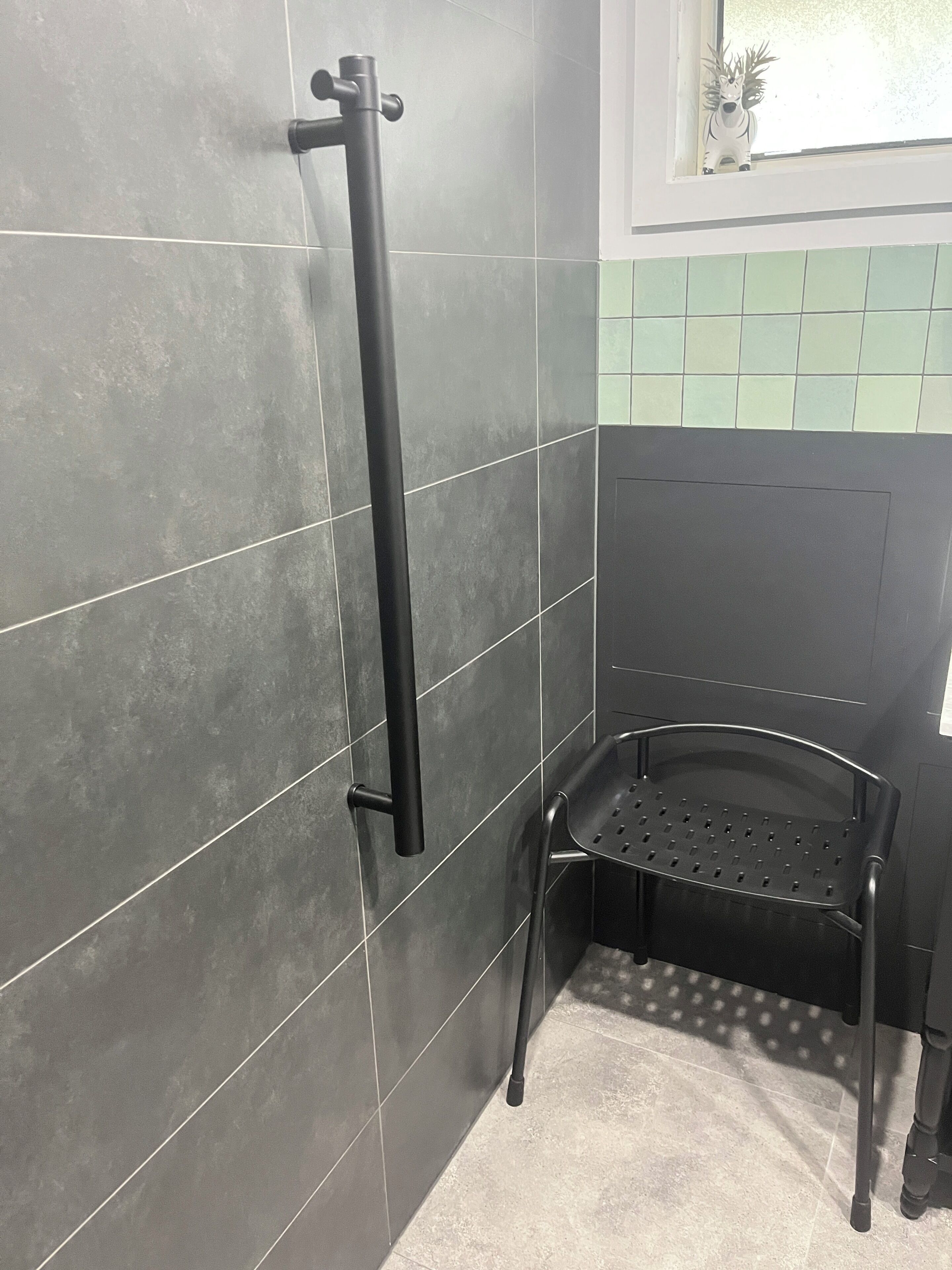 A bathroom with dark grey tiled walls, a black grab bar, a black chair, and a window with a view of trees.