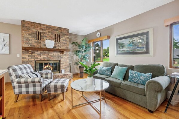 A living room with a brick fireplace, a sofa, a chair, a round table, and a large window.