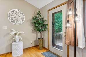 A hallway with a decorative wall piece, a potted plant, a white vase with flowers, and a door with a view of greenery outside.