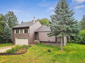 A two-story house with a garage, a front yard with a lawn and a flower bed, and a large evergreen tree.
